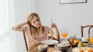 Teen girl in striped shirt at breakfast table with fruits, bread, and juice. Casual morning setting.